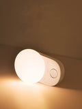 White USB rechargeable sensor night light with warm LED glow, shown on a bedroom nightstand.