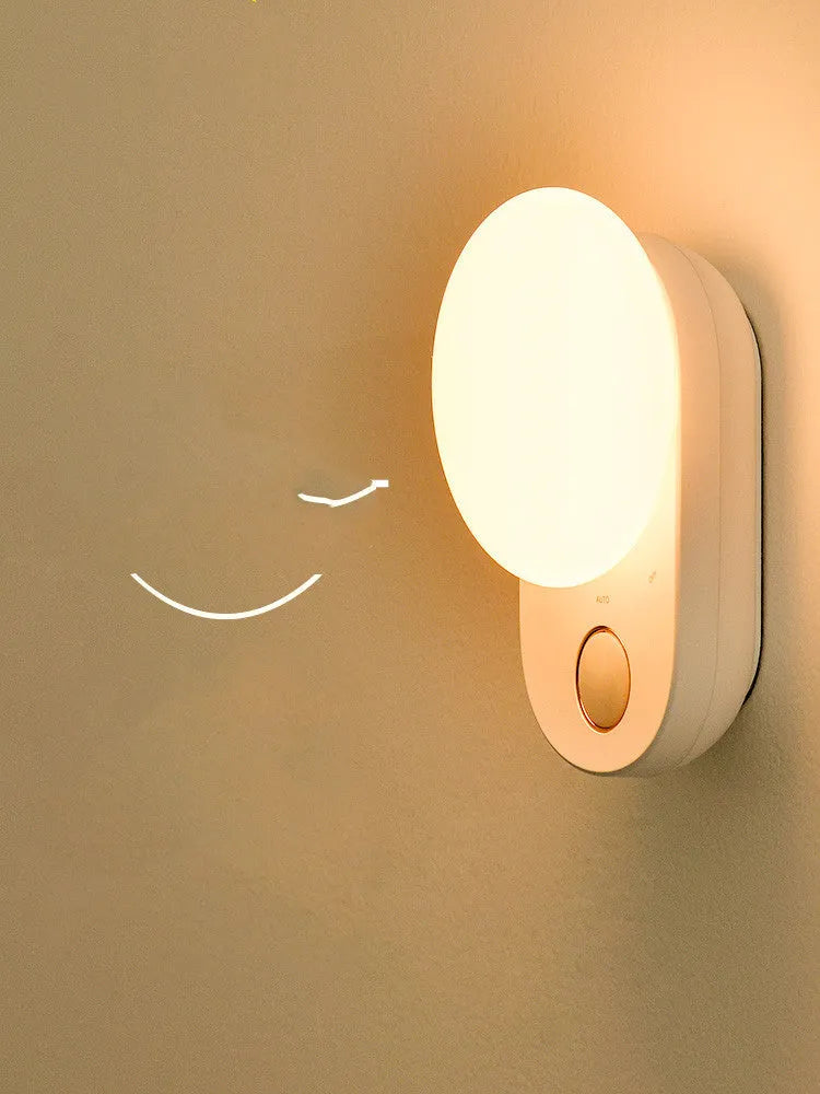 White USB rechargeable sensor night light with warm LED glow, shown on a bedroom nightstand.