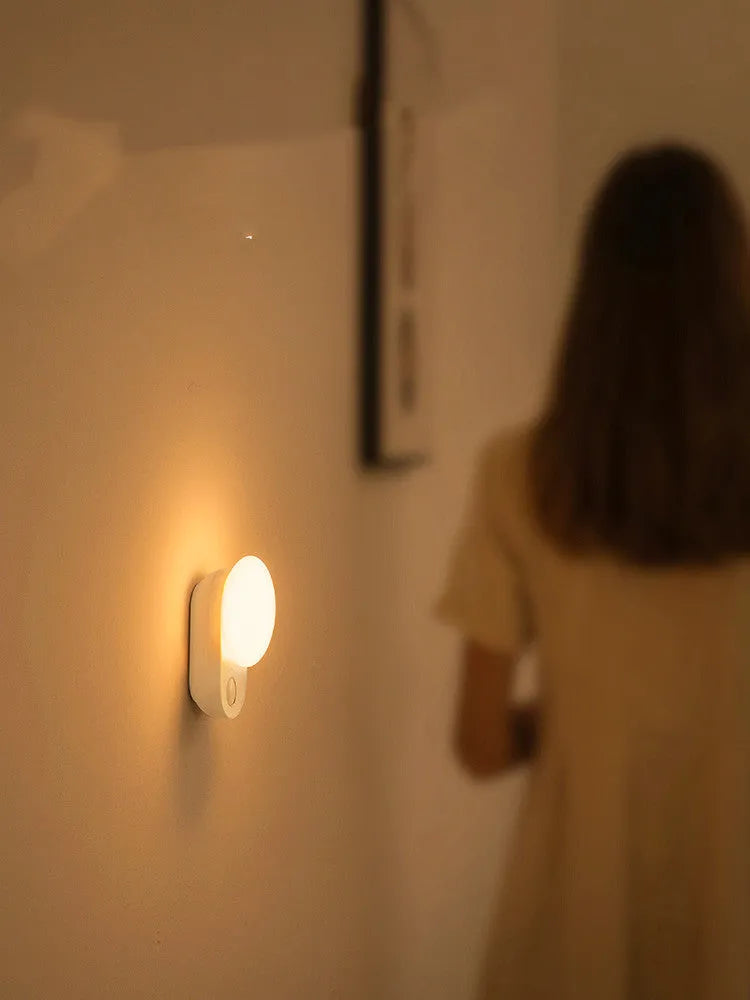 White USB rechargeable sensor night light with warm LED glow, shown on a bedroom nightstand.