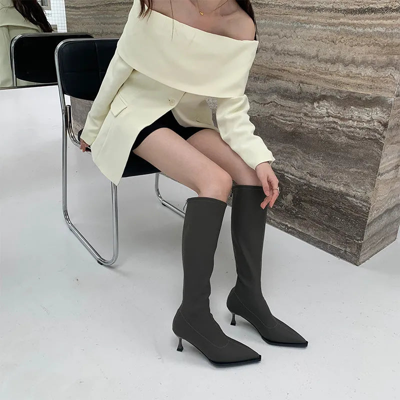 Black pointed toe below-the-knee boots on a woman, showing the back zipper and stiletto heel.