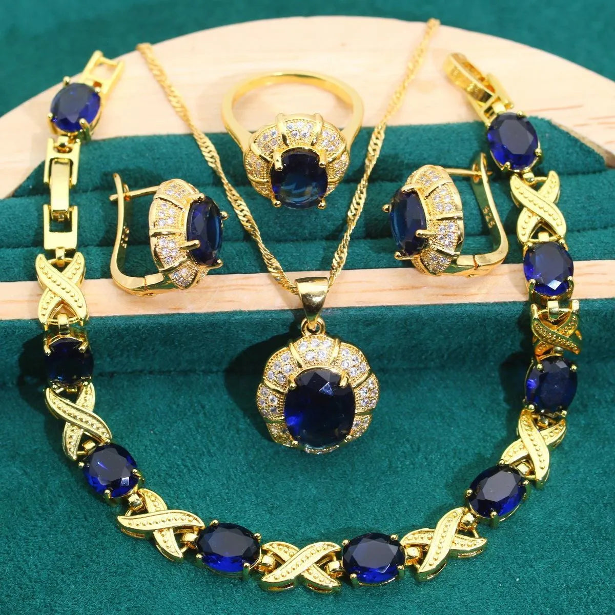 Gold plated necklace, earrings, ring, and bracelet set with blue cubic zirconia stones on a grey background.