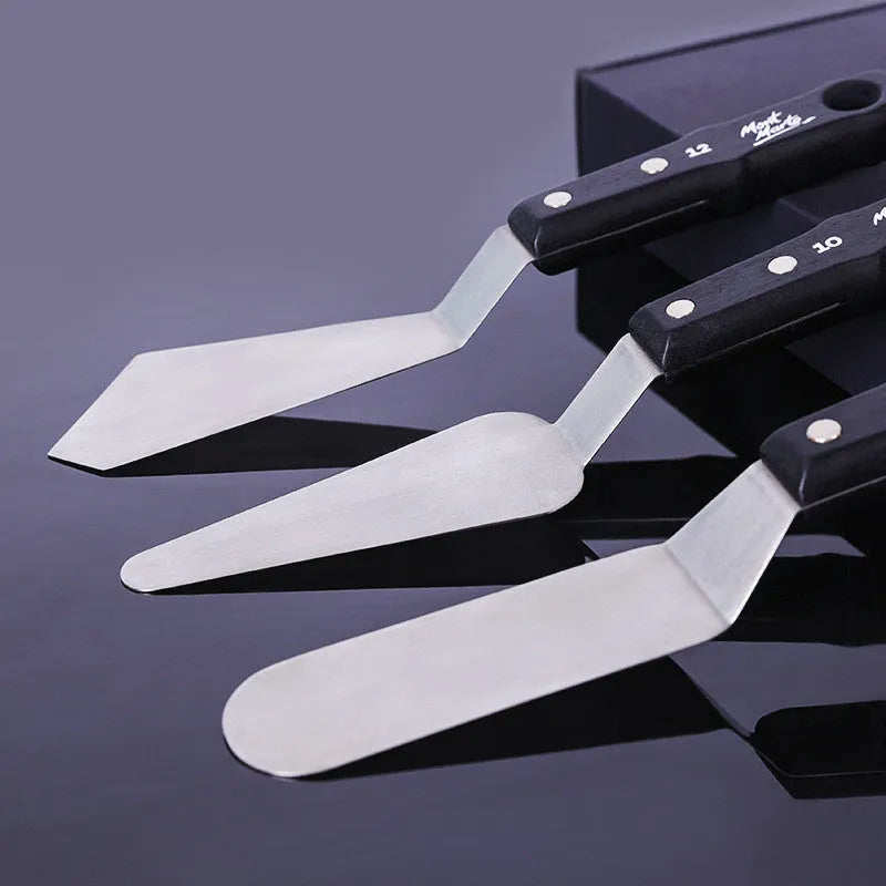 Three stainless steel artist palette knives with different blade sizes on a wooden table.