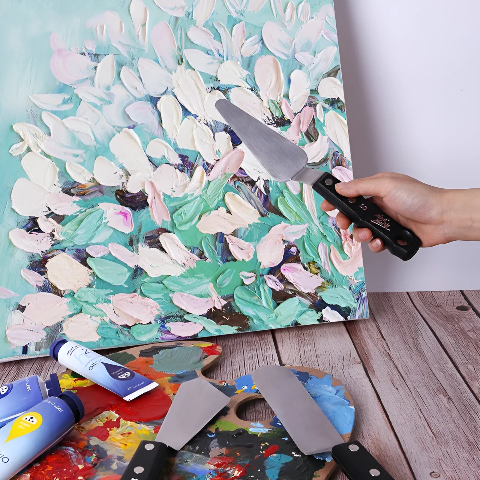 Three stainless steel artist palette knives with different blade sizes on a wooden table.