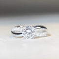 A silver moissanite ring with a 1 carat stone in a four-claw setting and a twisted geometric band.