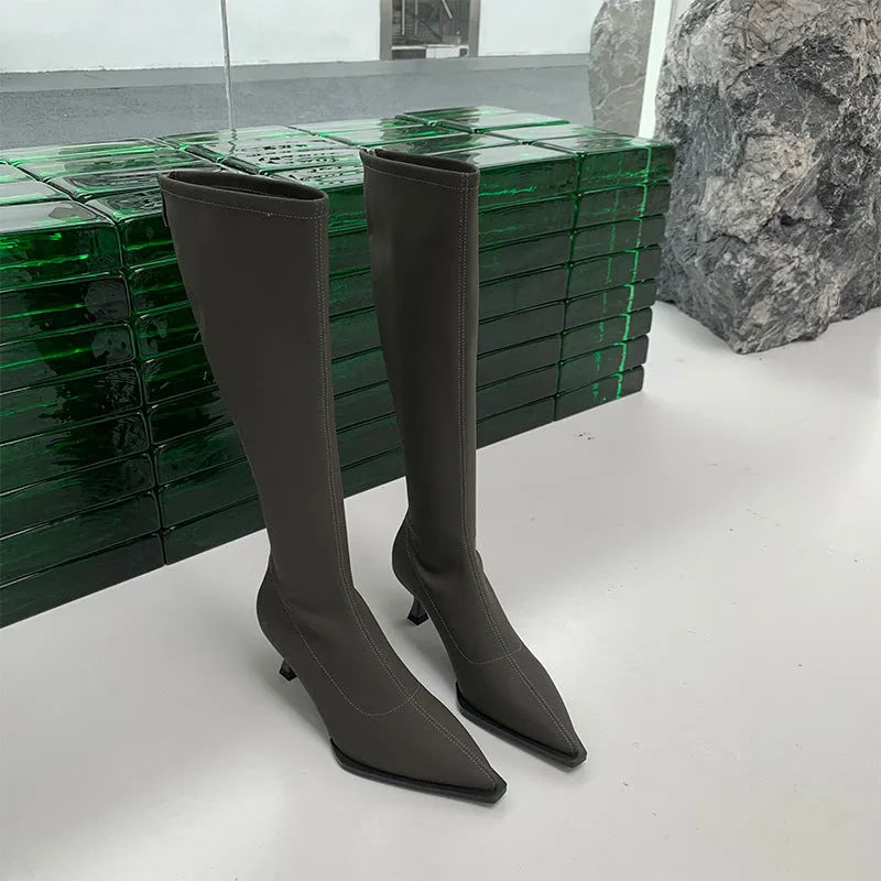Black pointed toe below-the-knee boots on a woman, showing the back zipper and stiletto heel.