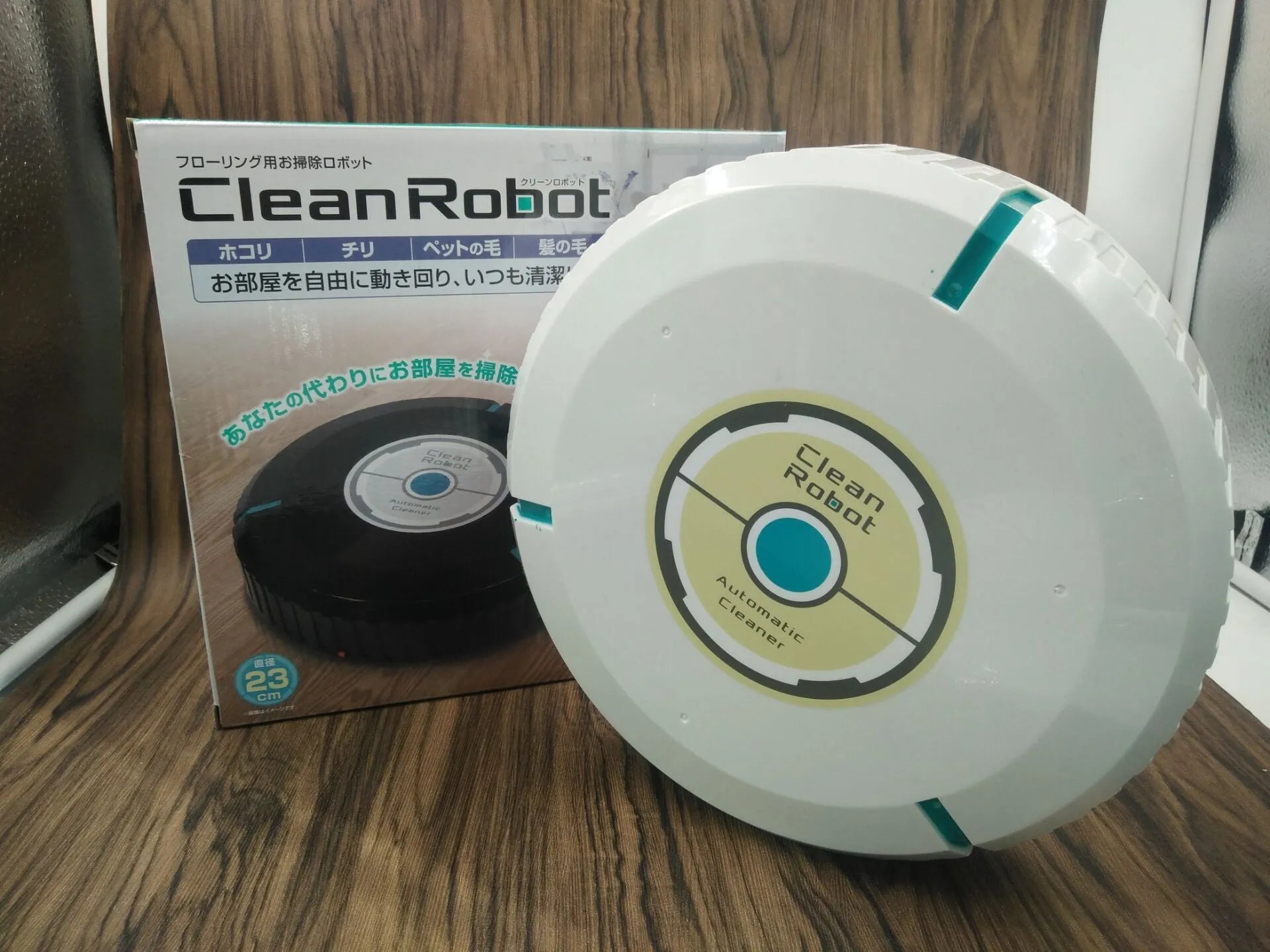 Black automatic sweeping robot on a wooden floor next to its packaging.