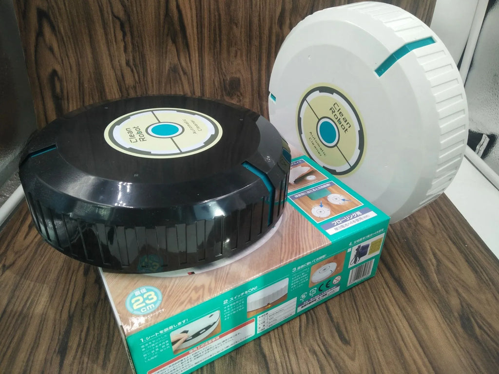 Black automatic sweeping robot on a wooden floor next to its packaging.