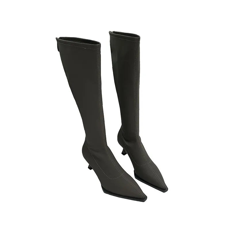 Black pointed toe below-the-knee boots on a woman, showing the back zipper and stiletto heel.