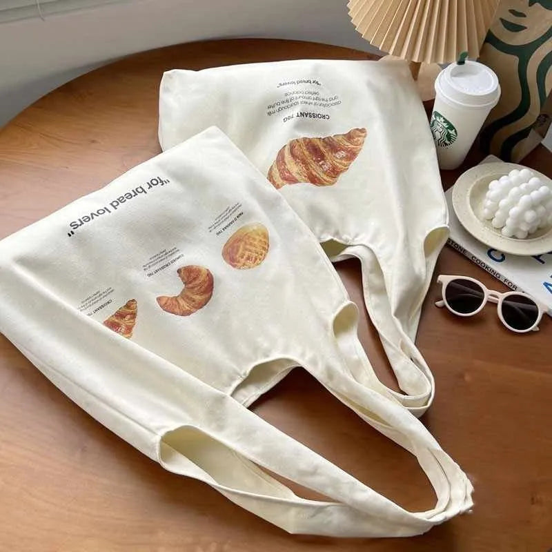 Beige canvas bag with bread patterns including croissant design, showing one-shoulder strap and structured form