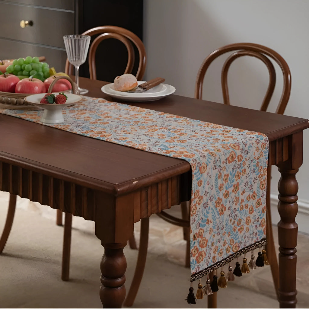 Beige jacquard table runner with long tassels and a woven flowers and leaves pattern draped over a table.