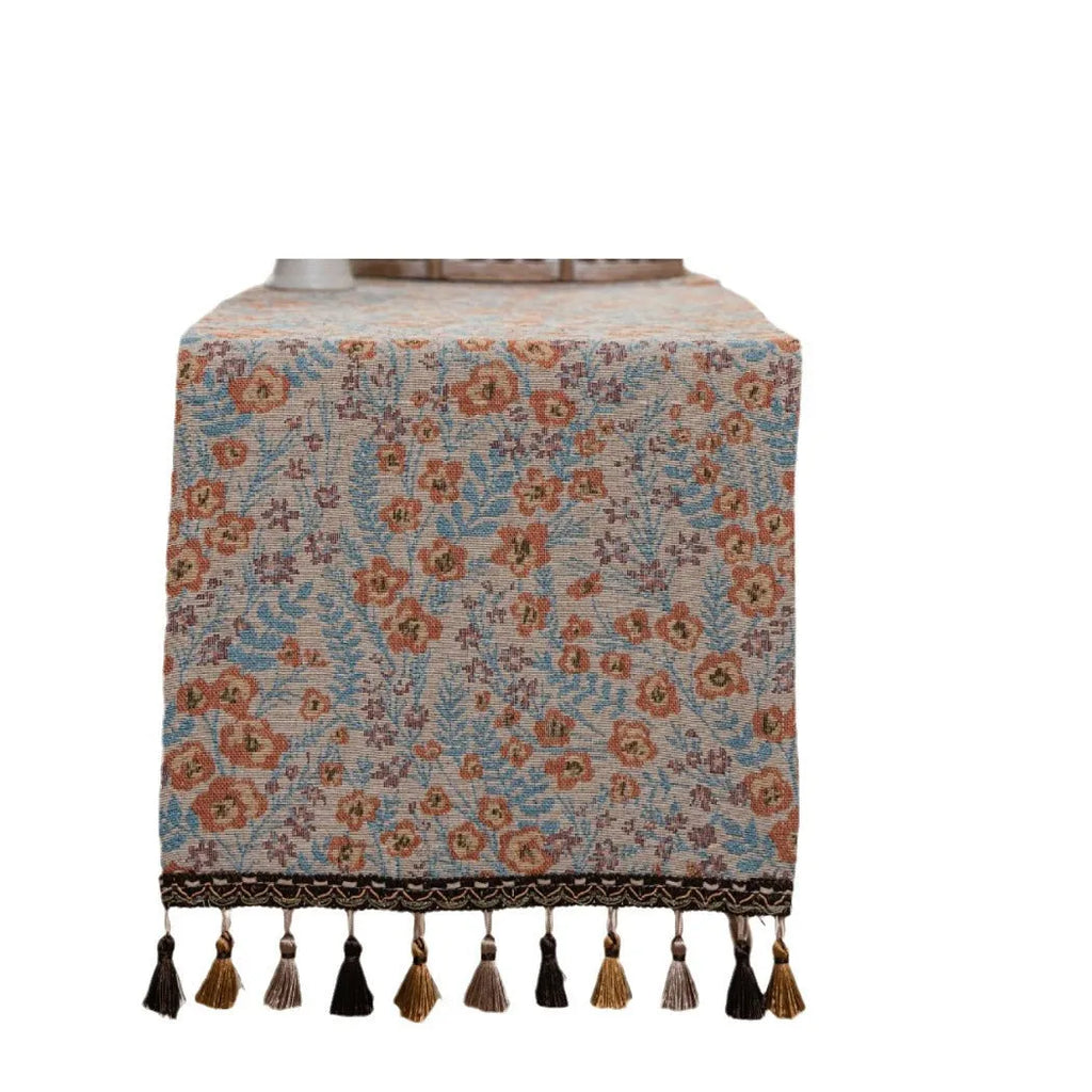 Beige jacquard table runner with long tassels and a woven flowers and leaves pattern draped over a table.