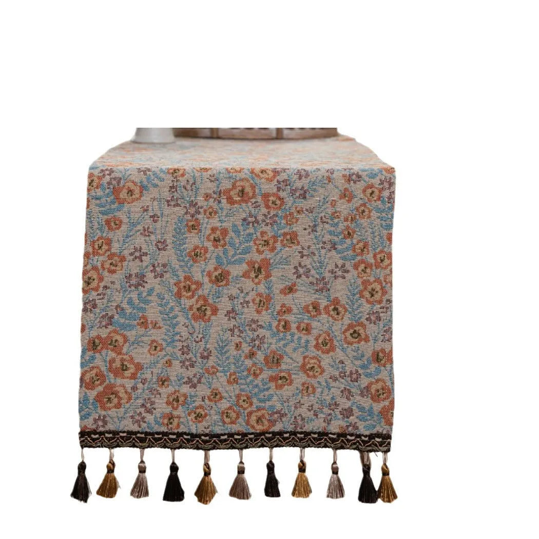Beige jacquard table runner with long tassels and a woven flowers and leaves pattern draped over a table.