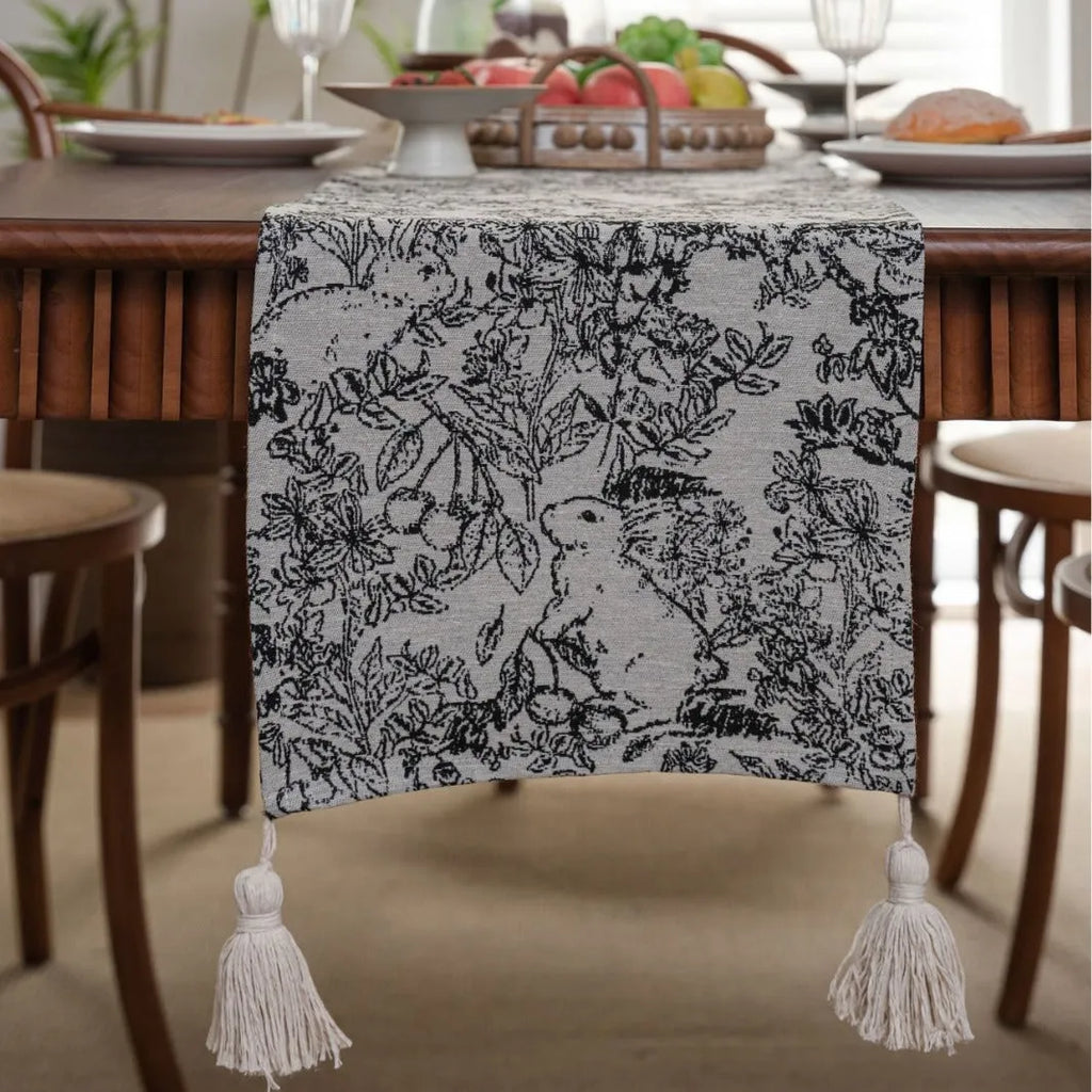 Beige jacquard table runner with long tassels and a woven flowers and leaves pattern draped over a table.