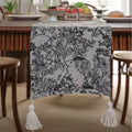 Beige jacquard table runner with long tassels and a woven flowers and leaves pattern draped over a table.