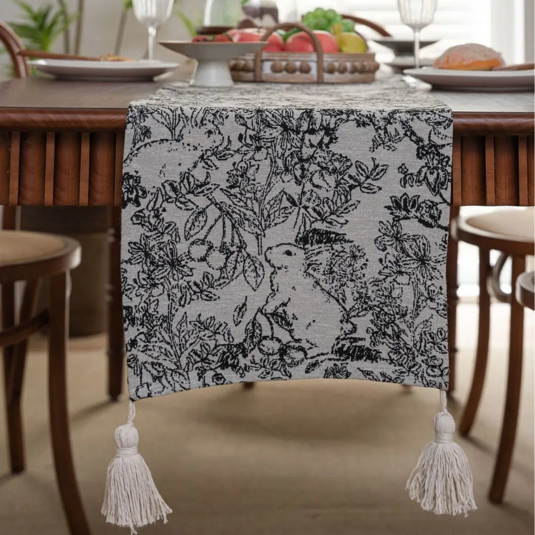 Beige jacquard table runner with long tassels and a woven flowers and leaves pattern draped over a table.