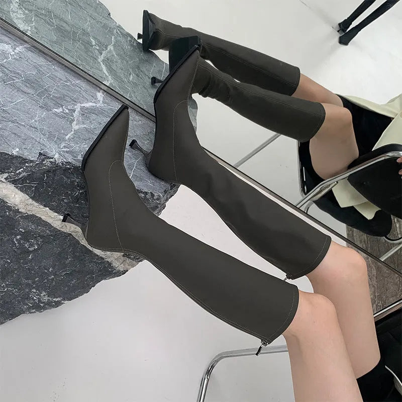 Black pointed toe below-the-knee boots on a woman, showing the back zipper and stiletto heel.