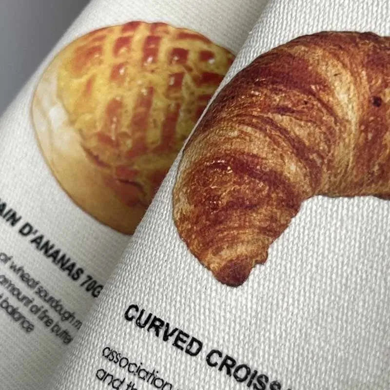 Beige canvas bag with bread patterns including croissant design, showing one-shoulder strap and structured form