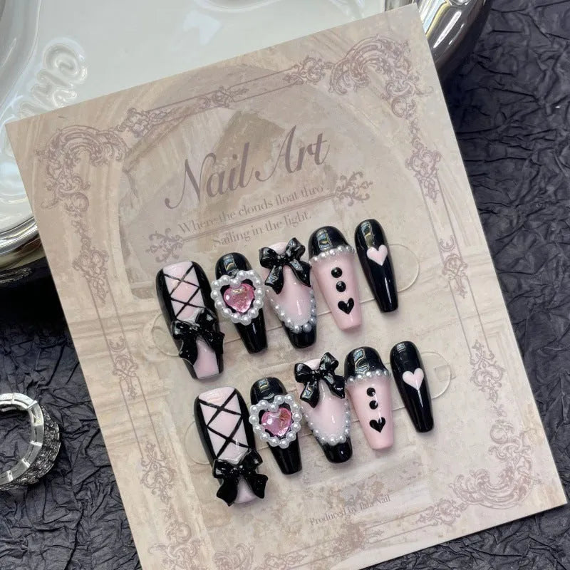 Hand-painted black artificial nails with pink hearts and a bow design, showing mid-length cat's eye style.